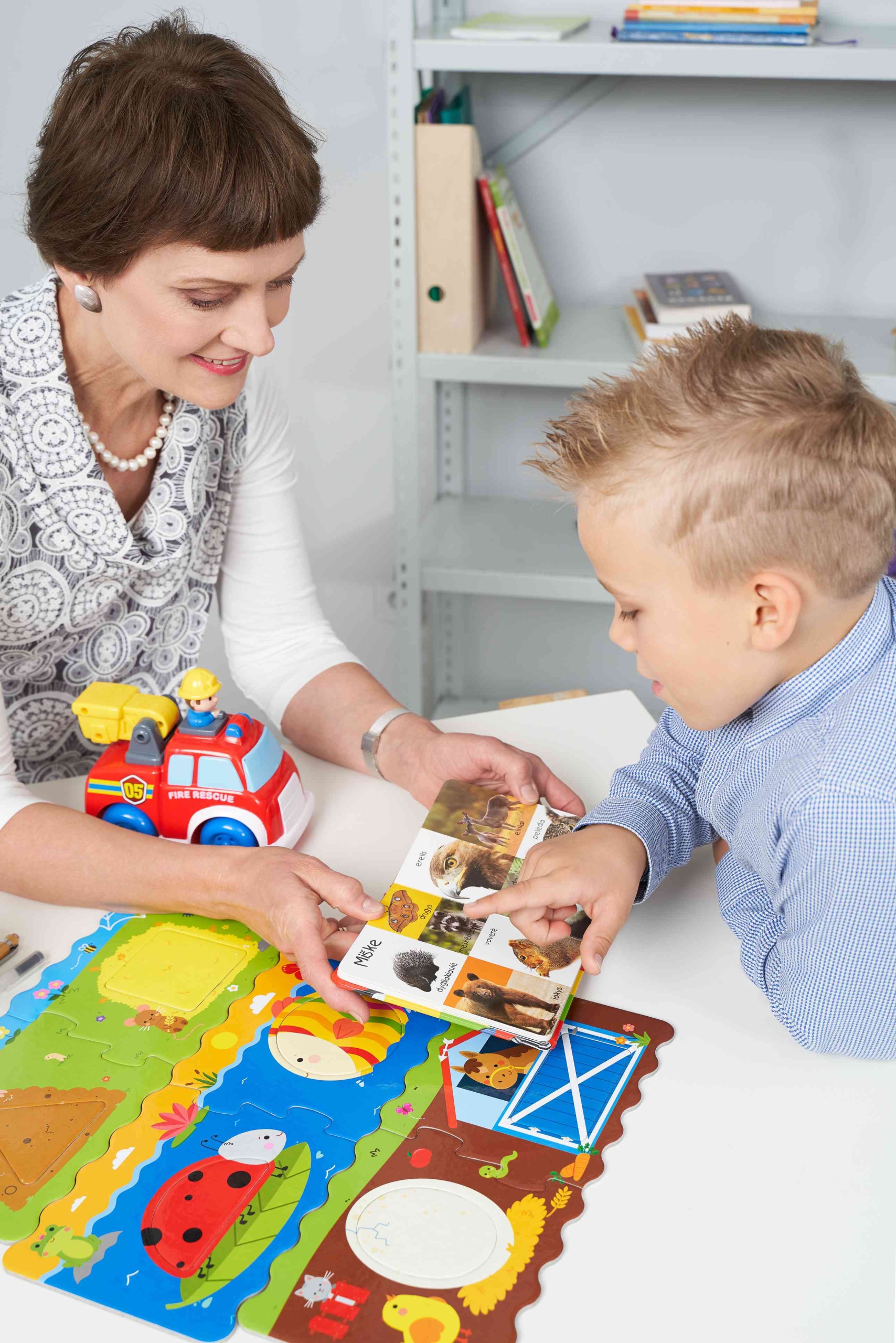 Logopedė Gražina Tumienė mokina vaiką logopedinio užsiėmimo metu. Rodomos kortelės ir lavinami vaiko motoriniai ir vizualiniai įgūdžiai.